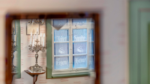 Fenster mit einer Gardine, mit dem GdF-Slogan Jeder Familie ihr Eigenheim, daneben ein Kerzenleuchter auf einem Regalbrett